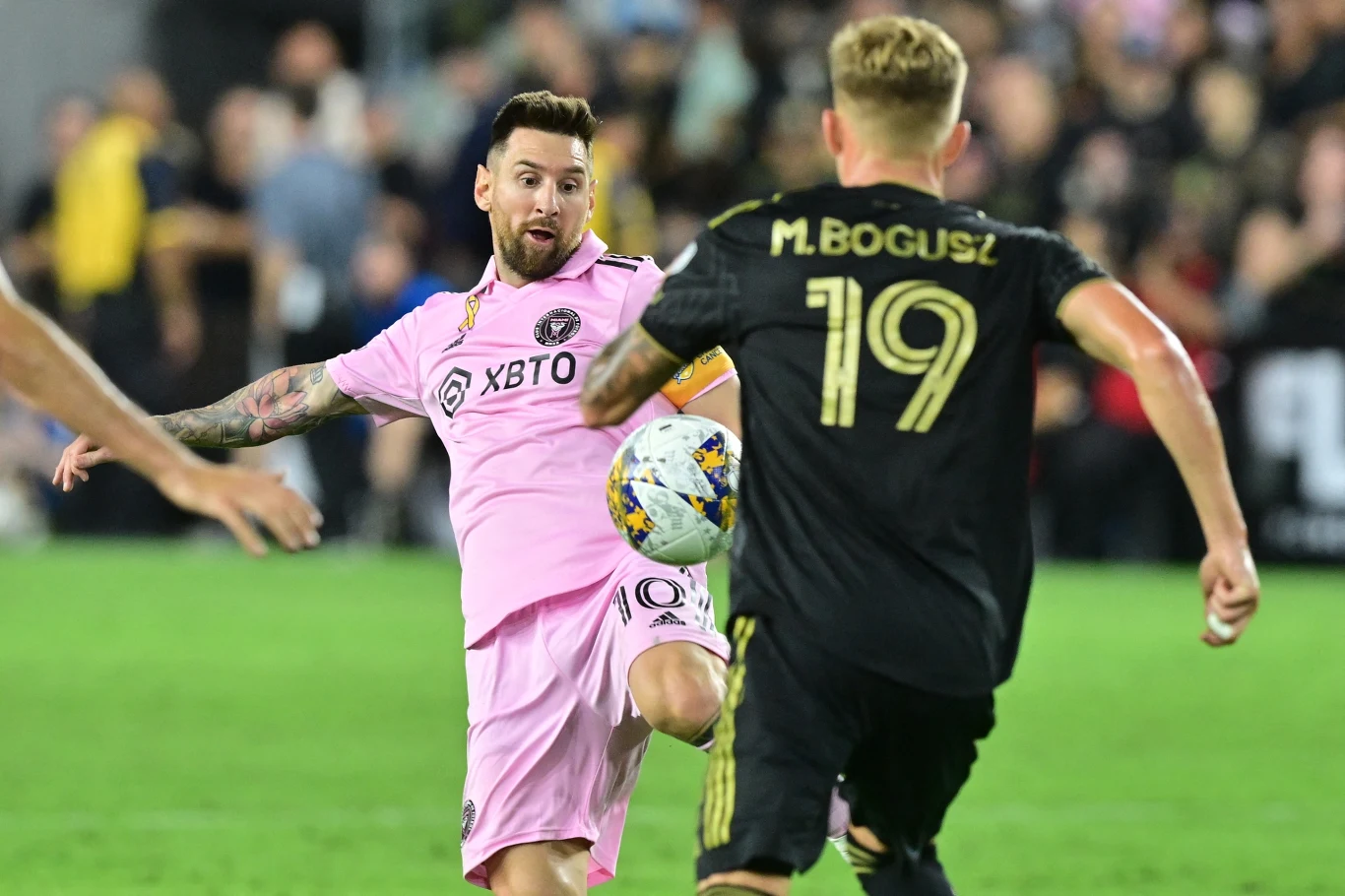 Dwóch piłkarzy walczy o piłkę podczas meczu na stadionie, jeden ubrany w różowy strój z logo sponsora, drugi w czarnym stroju z nazwiskiem oraz numerem na plecach, tłum kibiców w tle.