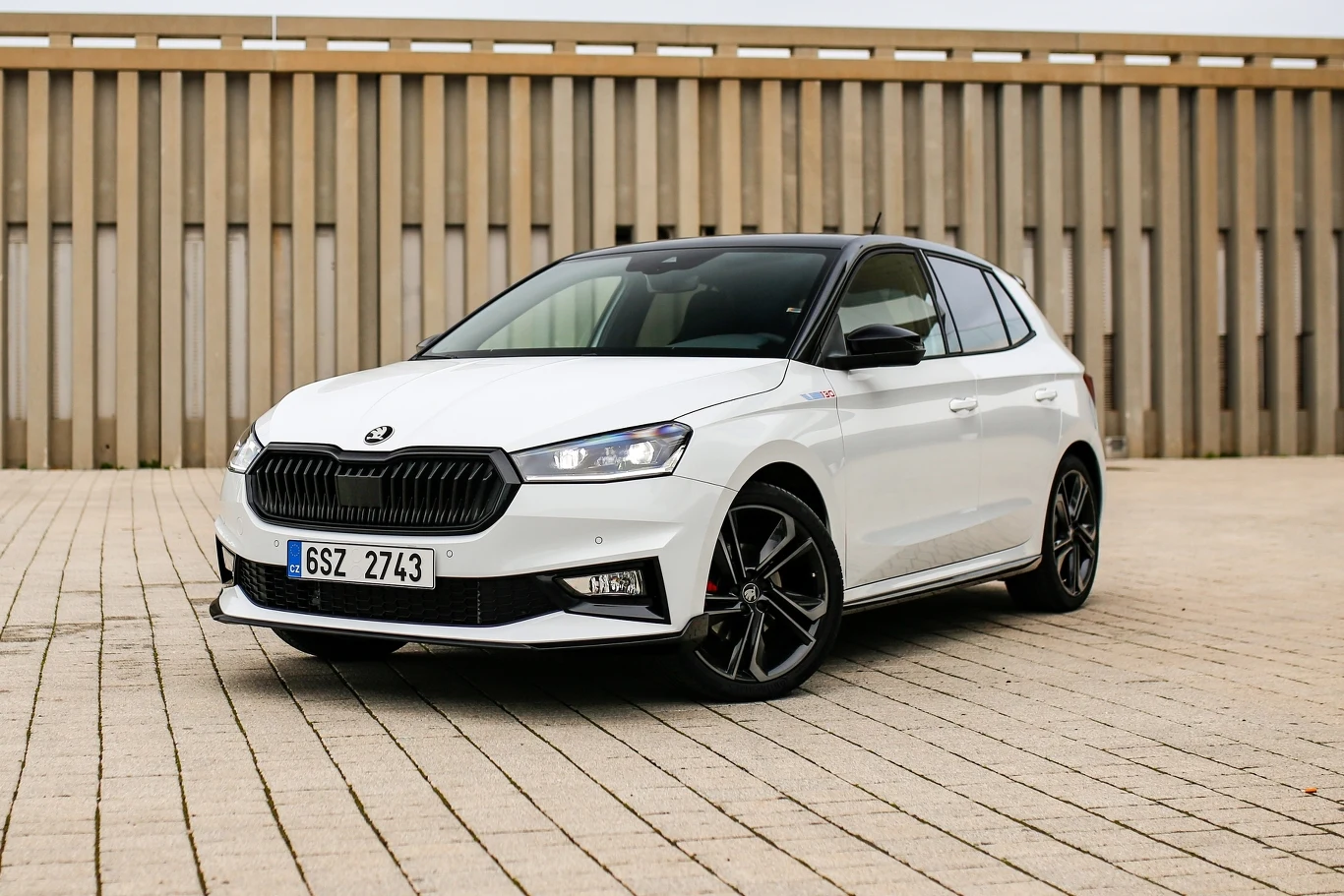 Biały samochód osobowy marki Skoda Fabia z czarnym dachem i czarnymi felgami stoi zaparkowany na jasnobrązowej kostce brukowej przed drewnianą nowoczesną ścianą.