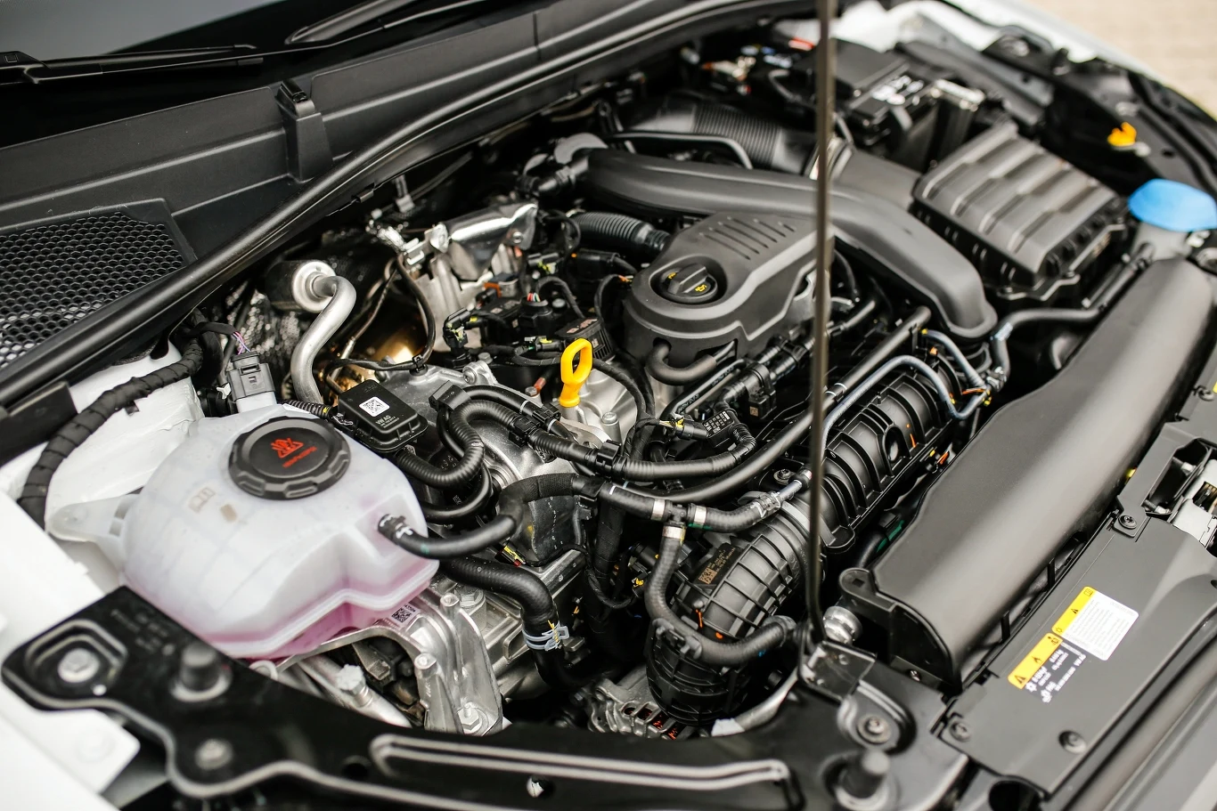 Komora silnika samochodowego z widocznymi komponentami, takimi jak blok silnika, zbiornik płynu chłodniczego, przewody i elementy systemu zasilania oraz osłony. Wszystkie części są czyste i dobrze widoczne.