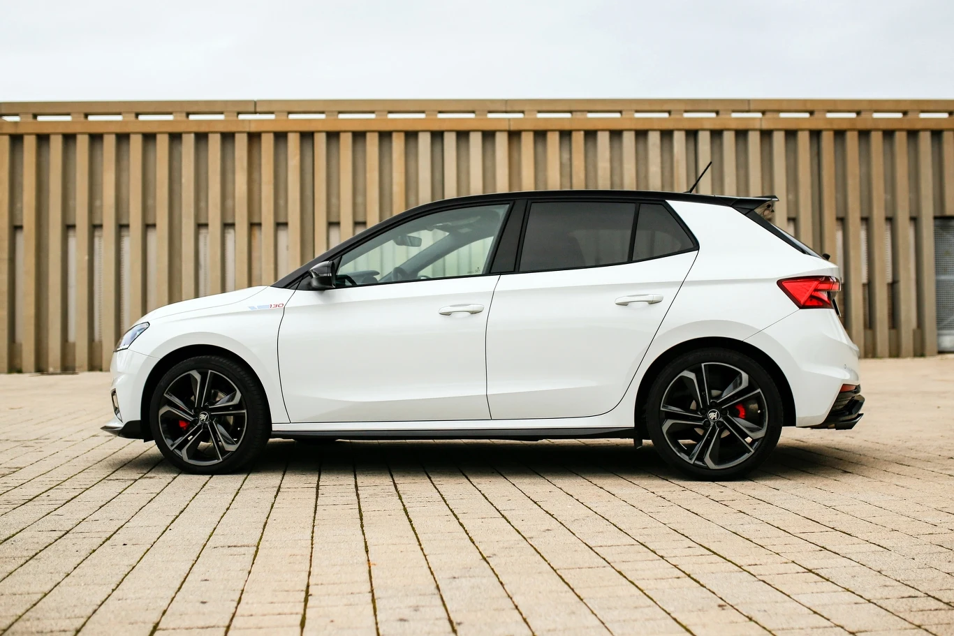 Biały samochód osobowy typu hatchback z czarnym dachem i detalami, ustawiony bokiem na tle betonowego placu oraz budynku z pionowymi elementami architektonicznymi. Sportowe felgi oraz nowoczesny wygląd auta.