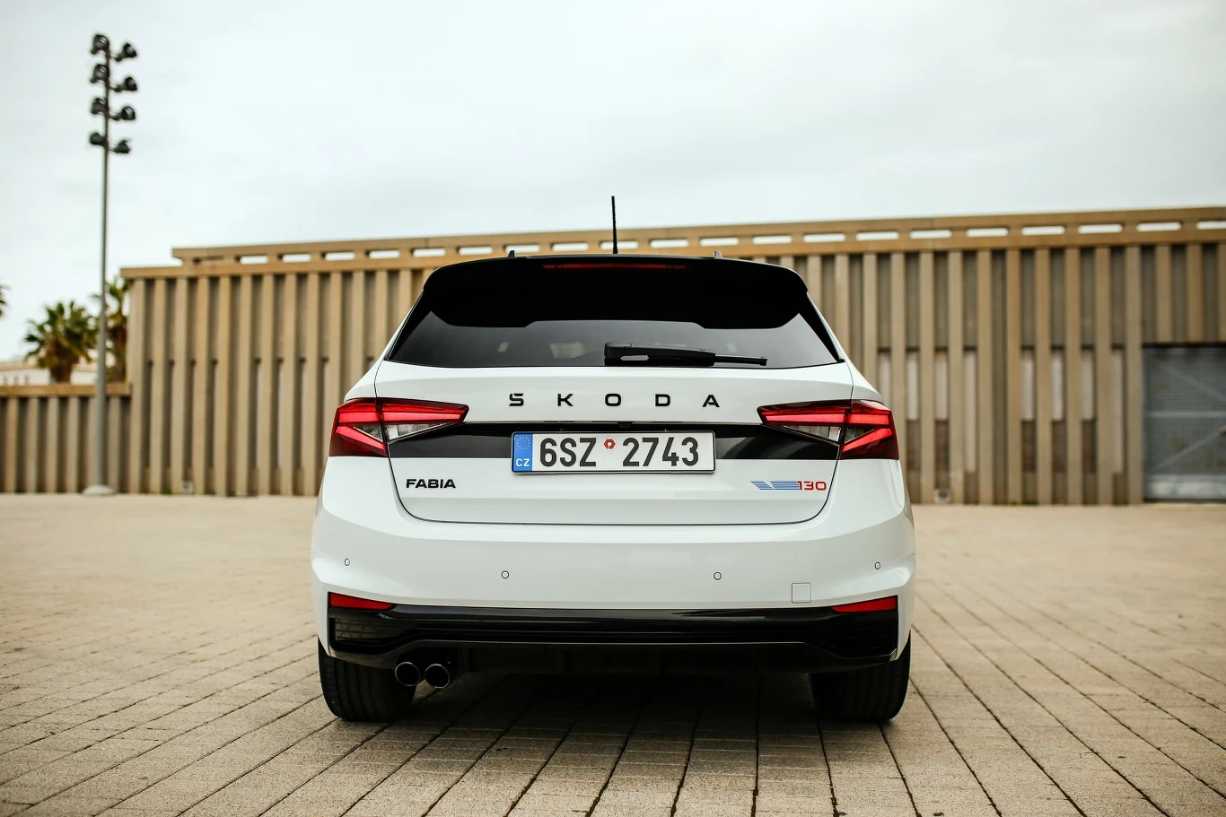 Biały samochód osobowy marki Skoda Fabia, widok od tyłu, stoi na pustym placu z kostki brukowej. W tle znajduje się nowoczesny budynek o pionowych przesłonach i jasno-szare niebo. Tylna szyba samochodu przyciemniona, tablica rejestracyjna czeska.