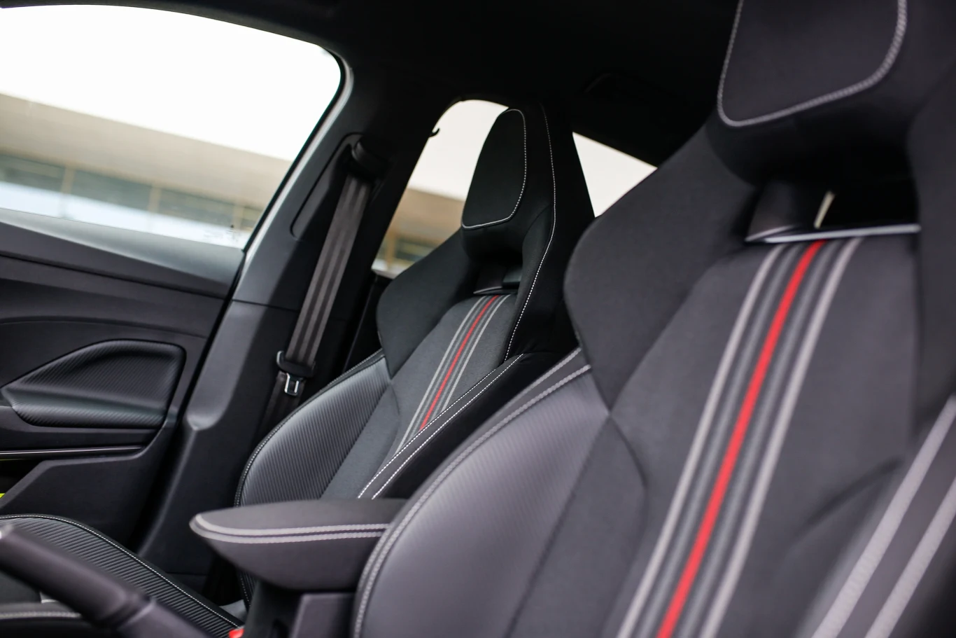 Fotele samochodowe z czarnej skóry z wyraźnymi czerwonymi i białymi przeszyciami w nowoczesnym wnętrzu auta. W tle widoczne fragmenty drzwi oraz elementy środkowego tunelu.