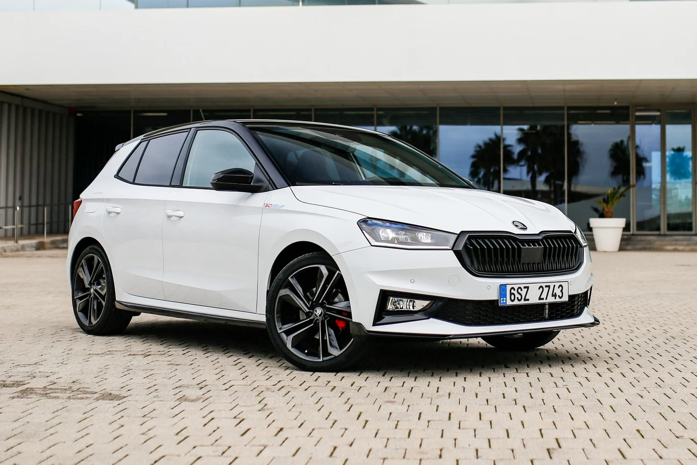 Biały samochód osobowy marki Škoda Fabia zaparkowany na placyku przed nowoczesnym budynkiem, wyraźnie widoczne sportowe felgi, czarne lusterka boczne oraz reflektory LED.