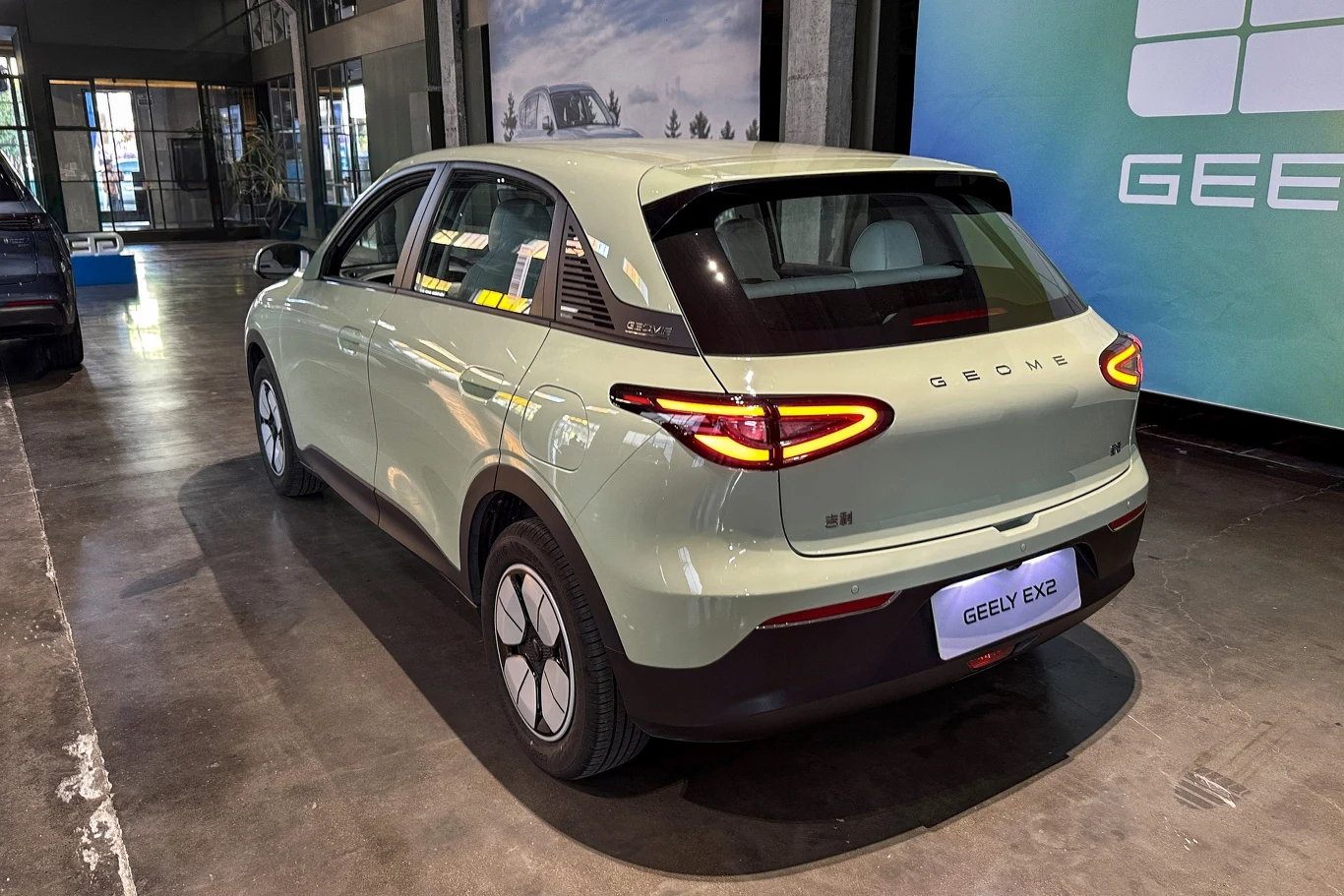 Nowoczesny samochód elektryczny Geely EX3 w jasnym kolorze, zaparkowany w przestronnym, dobrze oświetlonym wnętrzu budynku. W tle wyraźnie widoczne logo marki Geely na dużym ekranie oraz inne pojazdy.