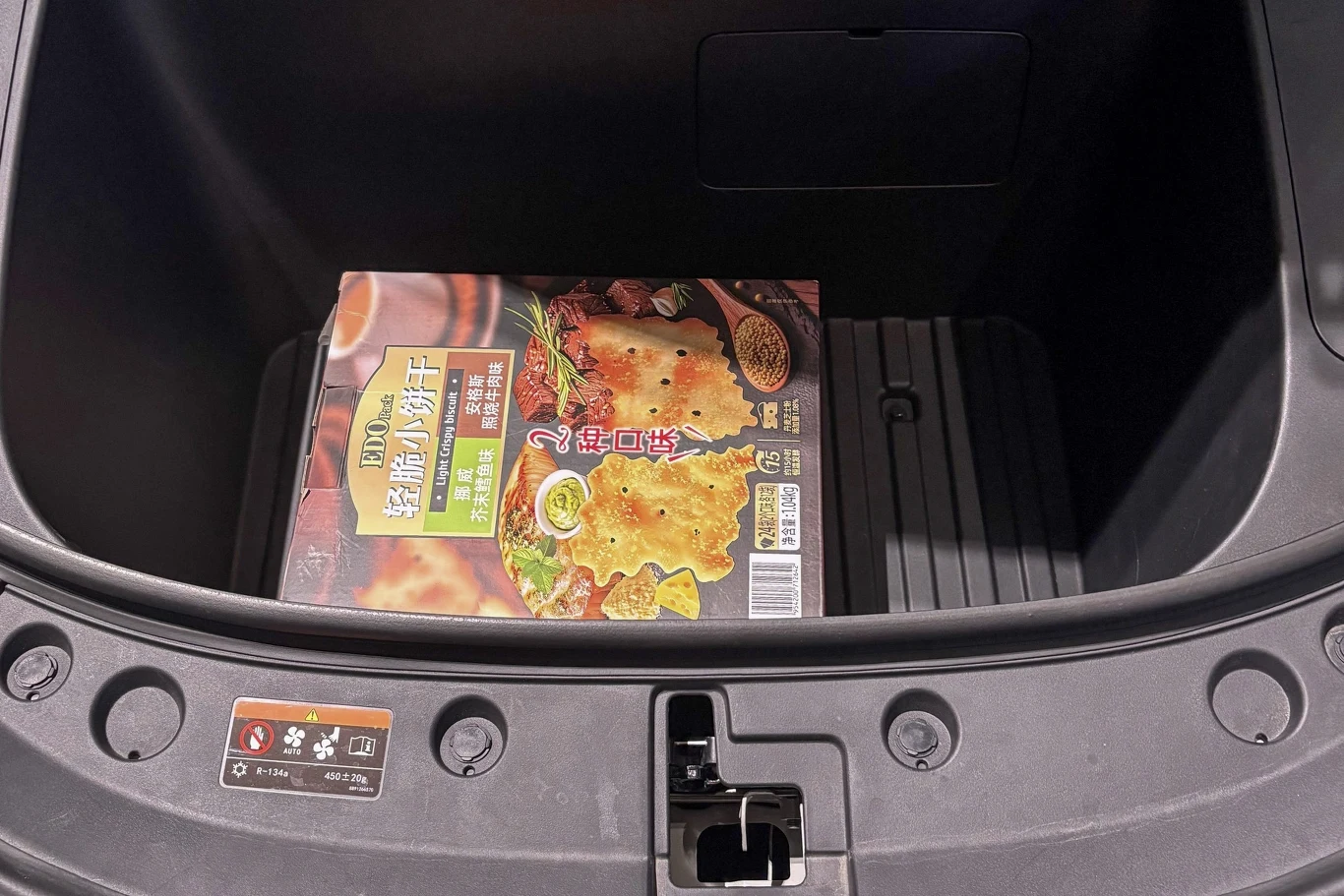 Kartonik z opakowaniem produktu spożywczego w języku chińskim umieszczony we wnętrzu bagażnika samochodu, otoczony ciemnymi plastikowymi ściankami bagażnika.