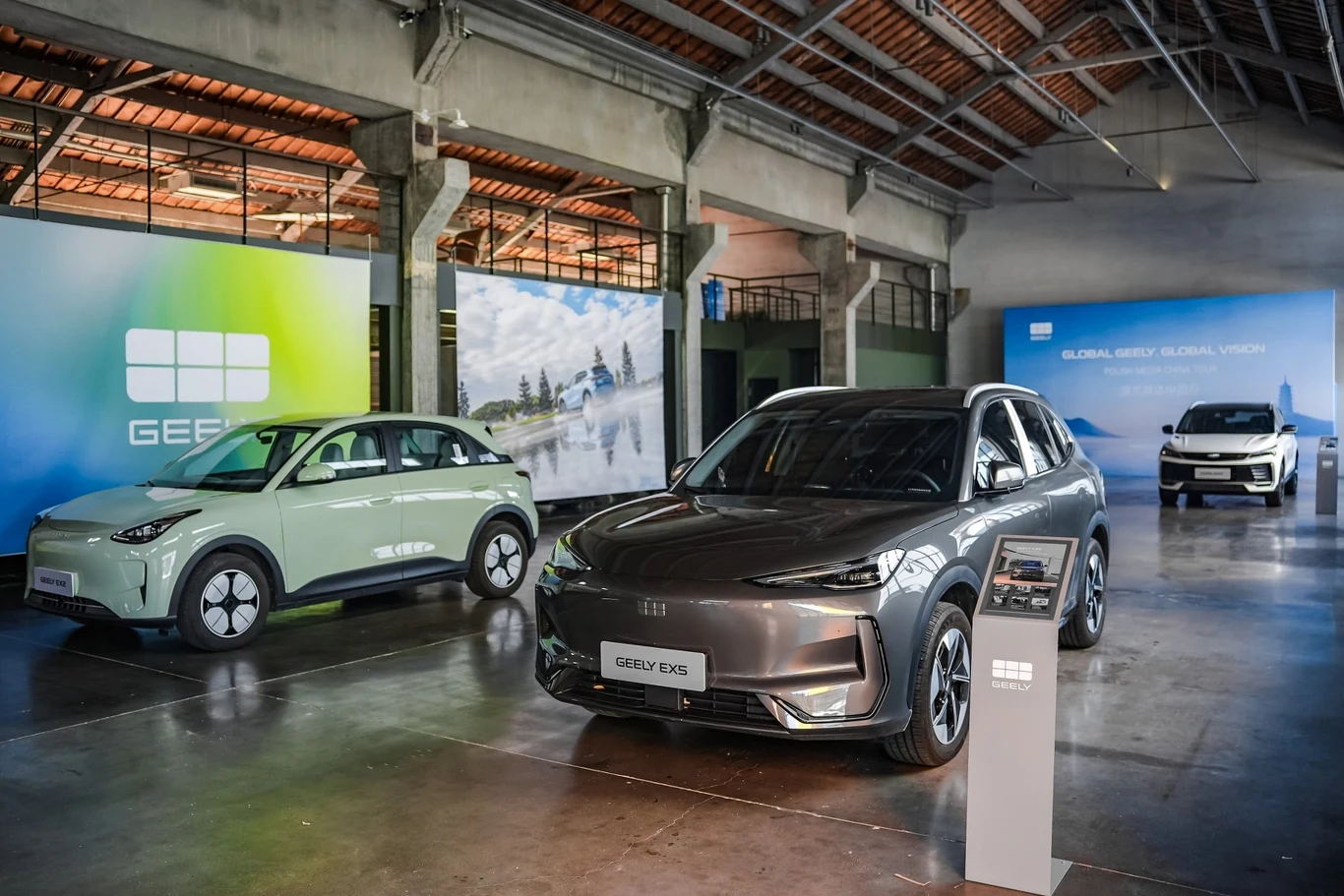 Nowoczesne samochody elektryczne ustawione w rzędzie w przestronnej hali wystawowej, z widocznymi planszami promocyjnymi marki Geely w tle.