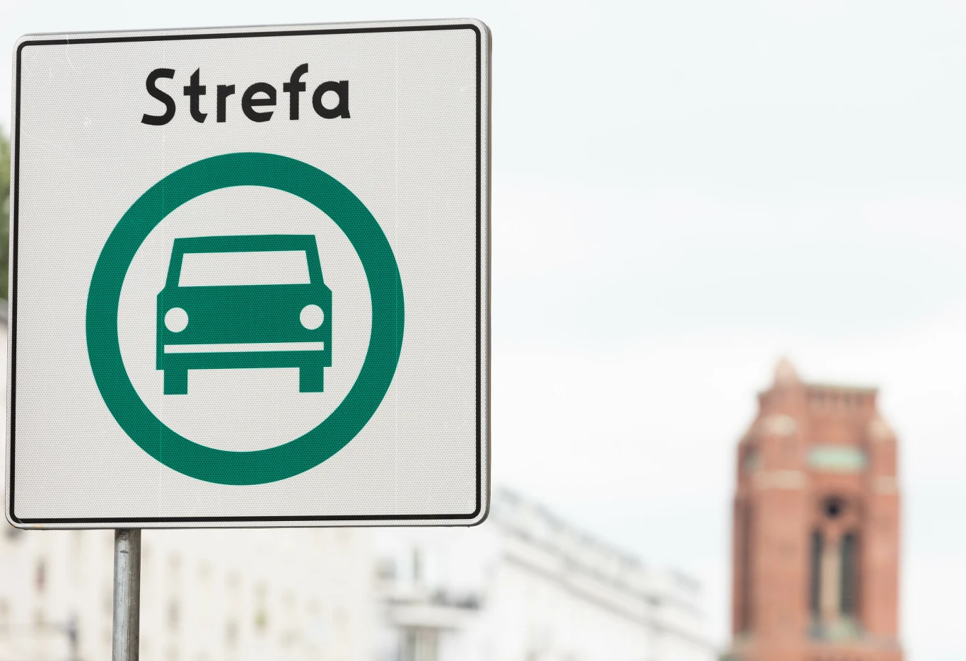 Kraków uruchamia Strefę Czystego Transportu Znak drogowy z napisem 'Strefa' oraz symbolem samochodu na tle miejskiej zabudowy z czerwoną wieżą w oddali.