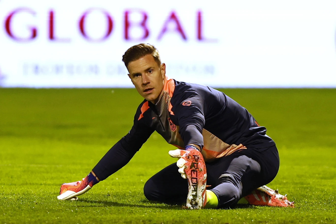 Bramkarz w stroju sportowym klęczy na murawie boiska piłkarskiego z jedną ręką wyciągniętą do przodu i skoncentrowaną miną, w tle rozmazany baner reklamowy.