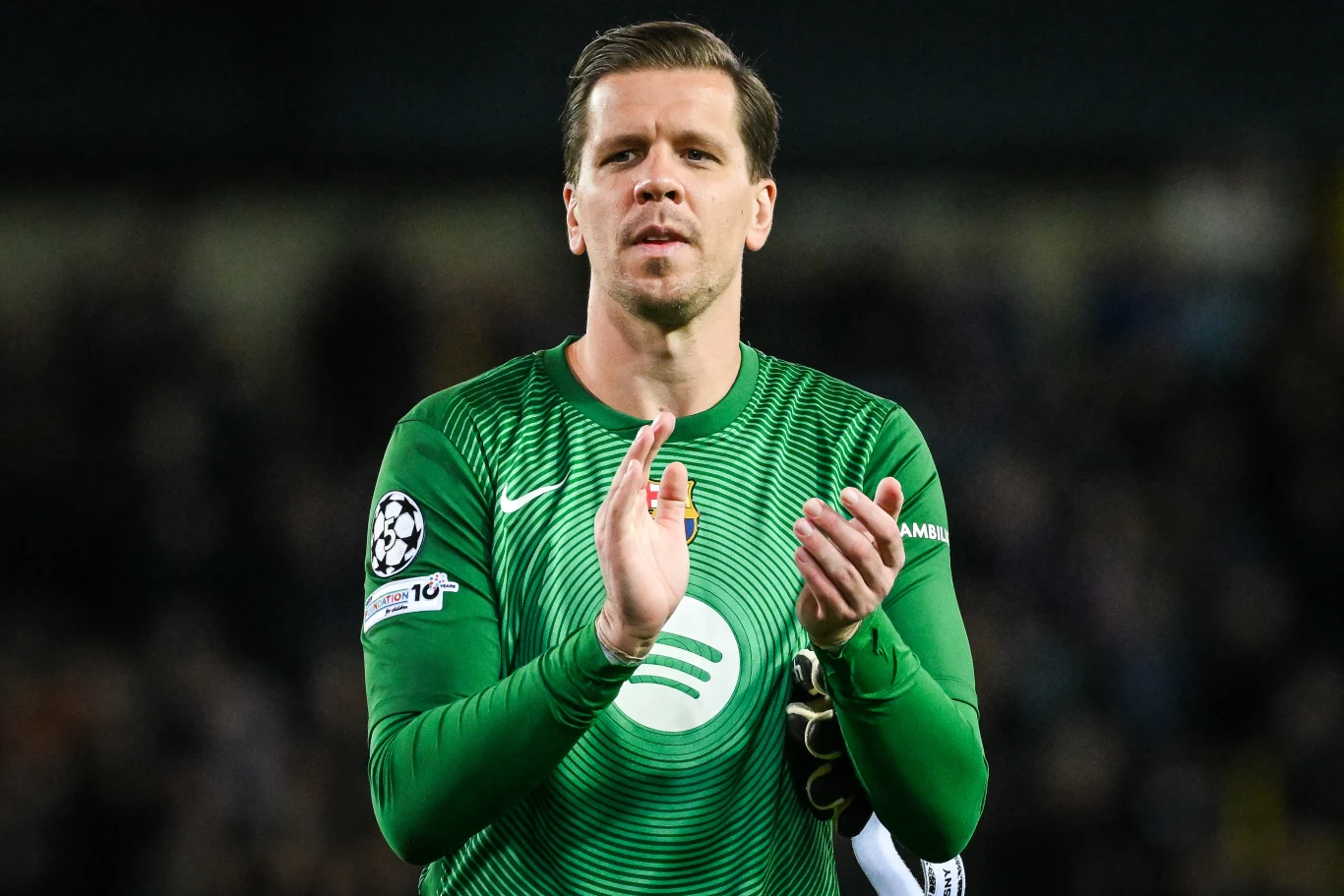 Bramkarz ubrany w zielony strój sportowy z logo klubu piłkarskiego i opaską na ramieniu, klaszcze w dłoniach, stojąc na tle zamazanego stadionu.