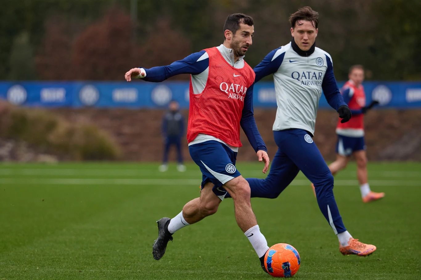 Dwóch piłkarzy w trakcie treningu na murawie, jeden z nich próbuje odebrać piłkę drugiemu, skupienie i sportowa rywalizacja widoczne na ich twarzach, tło stanowi rozmyta roślinność oraz banery reklamowe przy boisku.