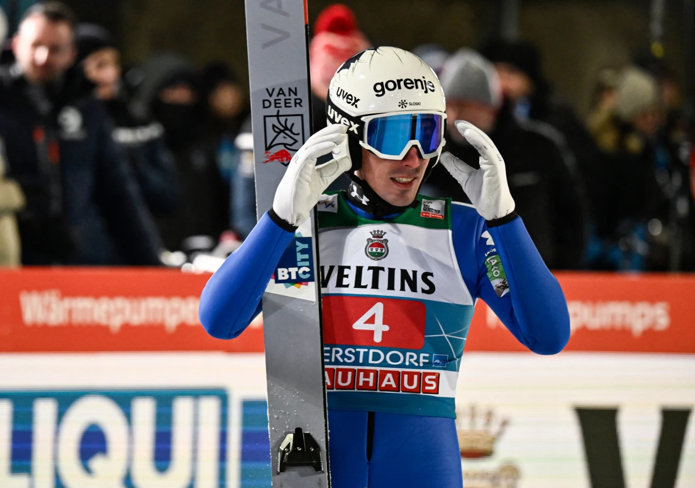 Sportowiec w kombinezonie narciarskim i kasku z goglami stoi na tle banerów reklamowych, trzymając narty obok siebie, otoczony przez innych ludzi w tle.
