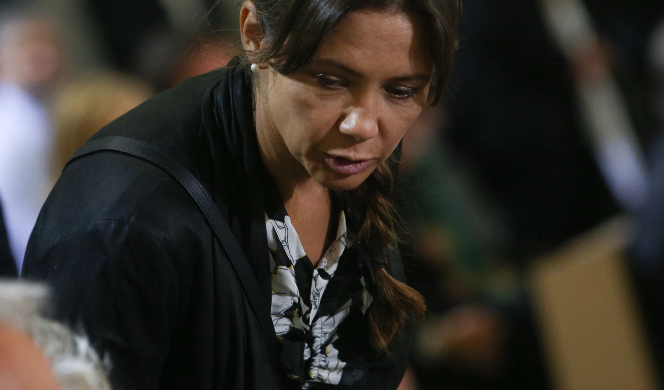 Kinga Rusin Kobieta o ciemnych włosach spiętych w warkocz, ubrana w czarny żakiet i wzorzystą bluzkę, pochylona nad rozmówcą w formalnym otoczeniu. W tle rozmazane sylwetki innych osób.