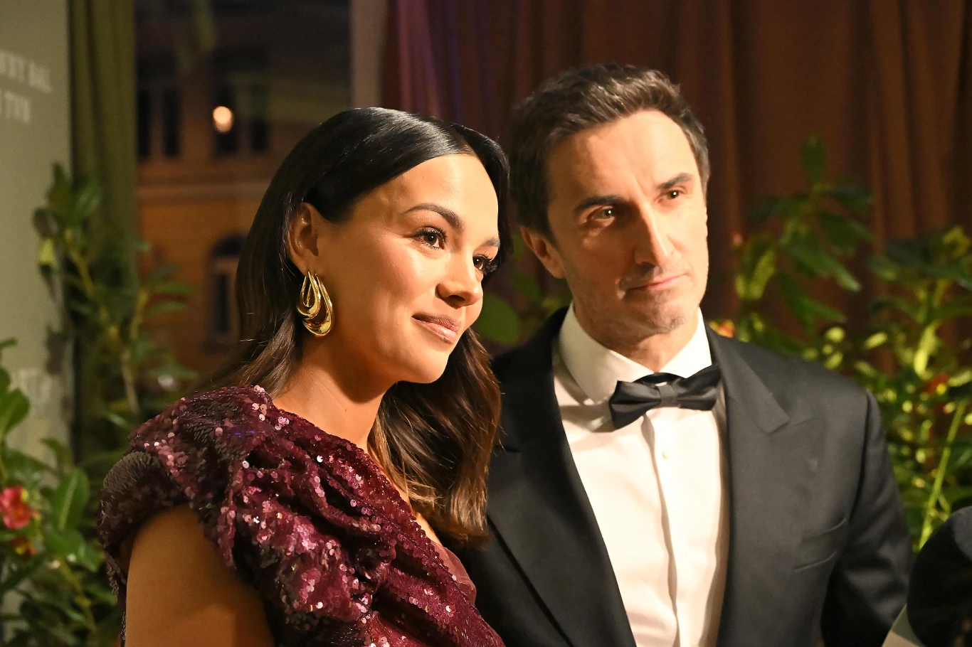 Paulina Krupińska-Karpiel, Sebastian Karpiel-Bułecka Elegancka kobieta w wieczorowej sukni oraz mężczyzna w smokingu pozujący razem, otoczeni zielenią i ciepłym światłem podczas uroczystego wydarzenia.