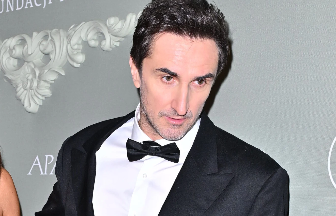 Sebastian Karpiel-Bułecka Mężczyzna ubrany w elegancki smoking i muchę, stojący na tle dekoracyjnej ściany z jasnymi ozdobnymi motywami.