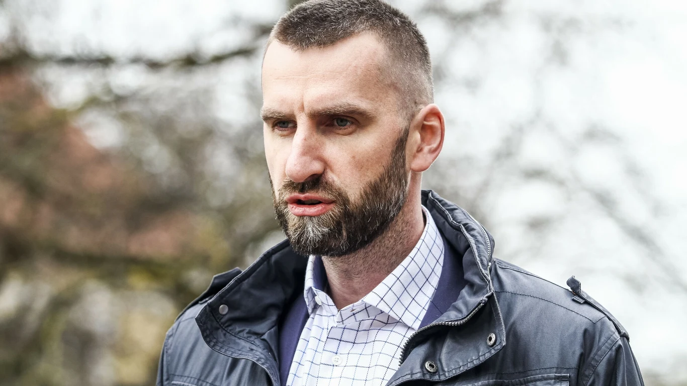 Mężczyzna z krótkimi włosami i brodą w kurtce skórzanej oraz koszuli w kratę patrzy przed siebie, tło rozmyte z gałęziami drzew.