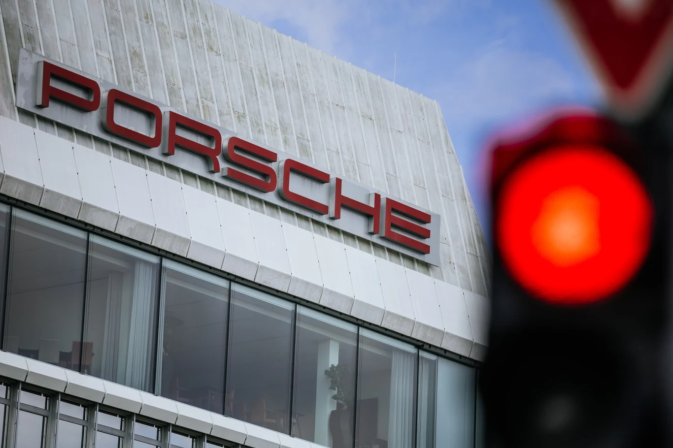 Nowoczesny budynek z dużymi oknami, na którym umieszczony jest charakterystyczny czerwony napis Porsche. Na pierwszym planie widoczna jest czerwona sygnalizacja świetlna.