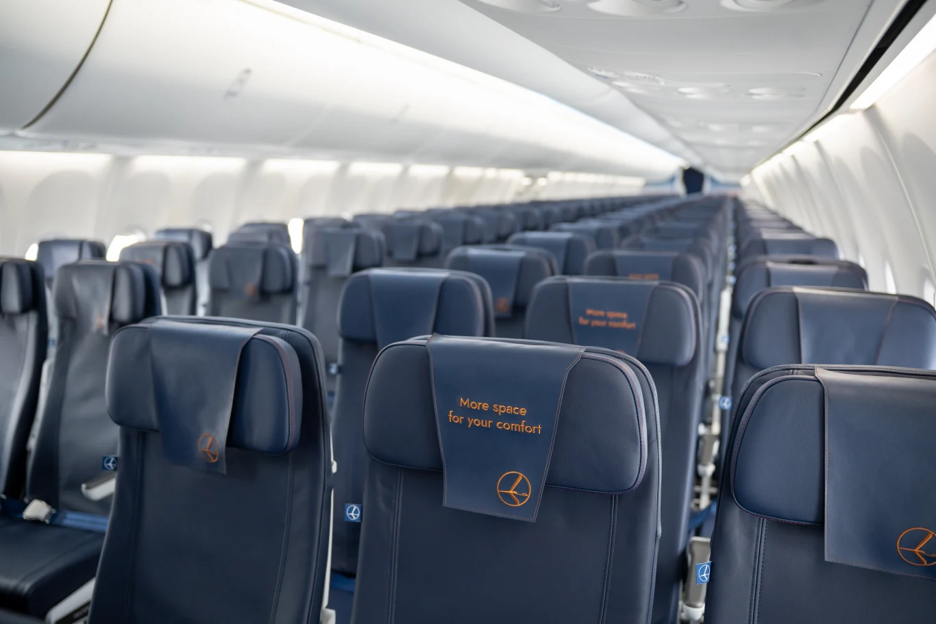Rzędy ciemnogranatowych, pustych foteli lotniczych z oparciami z wyhaftowanym napisem o większej przestrzeni dla komfortu podróży, dobrze oświetlone wnętrze kabiny pasażerskiej.