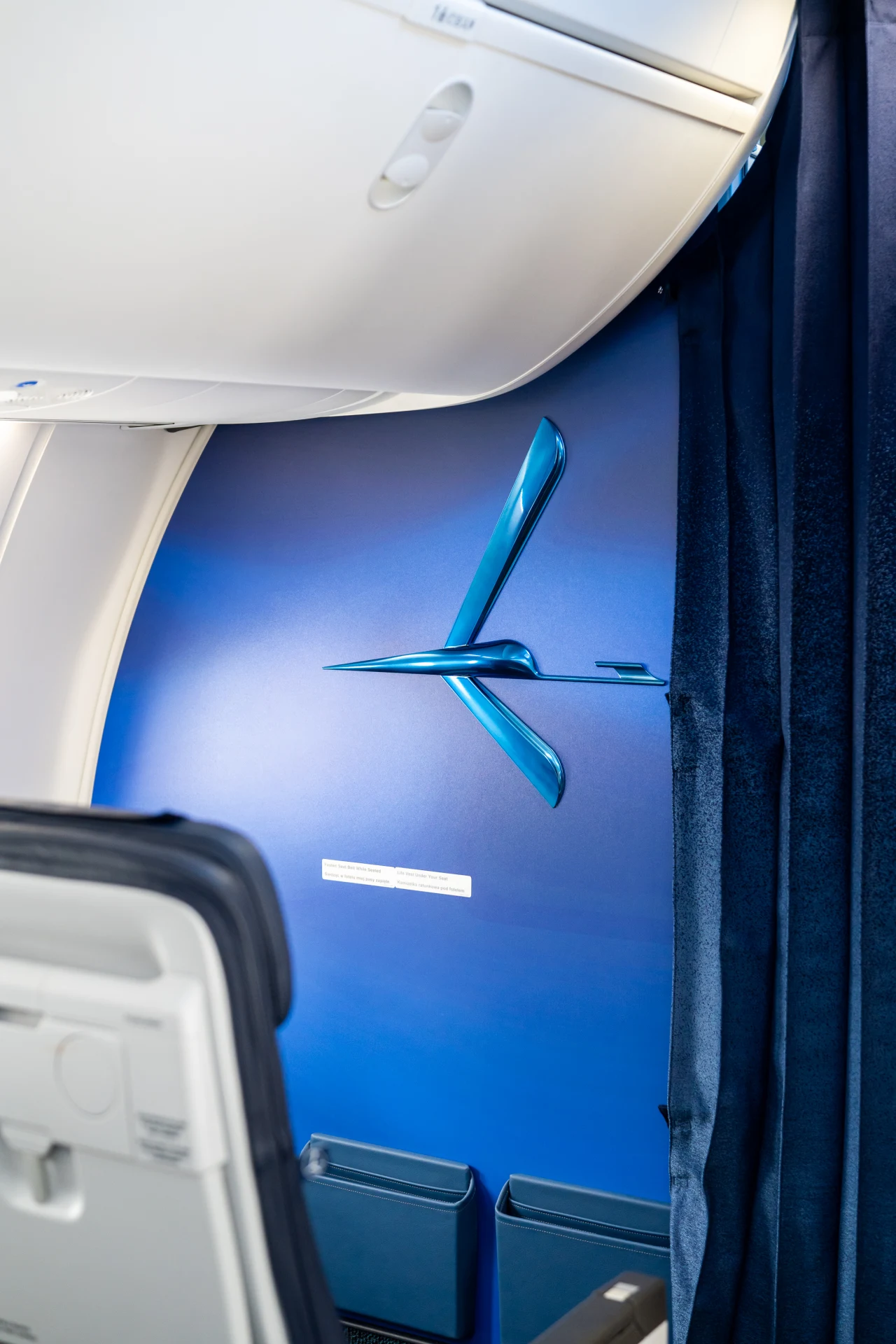 Fragment wnętrza samolotu pasażerskiego z widocznym niebieskim panelem z logo linii lotniczej oraz częścią fotela i zasłoną oddzielającą sekcję kabiny.