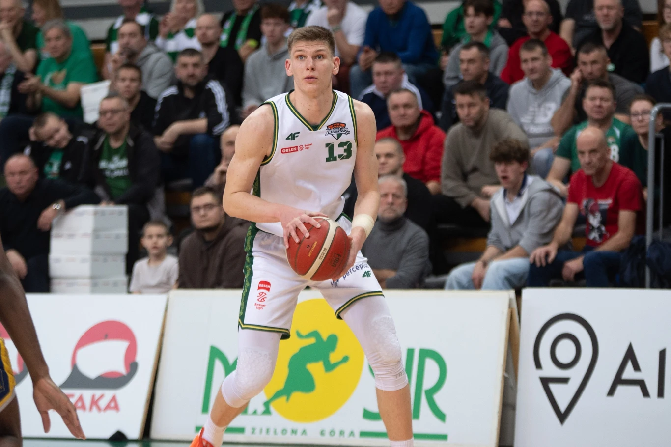 Koszykarz w białym stroju z numerem 13 trzyma piłkę oburącz, stojąc na boisku, w tle widoczna publiczność na trybunach skupiona na przebiegu meczu.