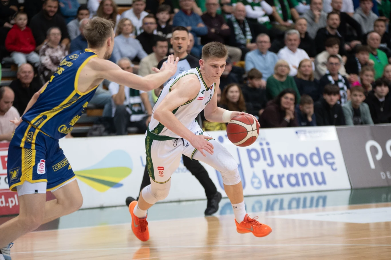 Koszykarz w białym stroju z piłką próbuje przedostać się obok obrońcy w niebiesko-żółtym stroju podczas meczu koszykówki. Na trybunach obserwuje wydarzenie licznie zgromadzona publiczność.