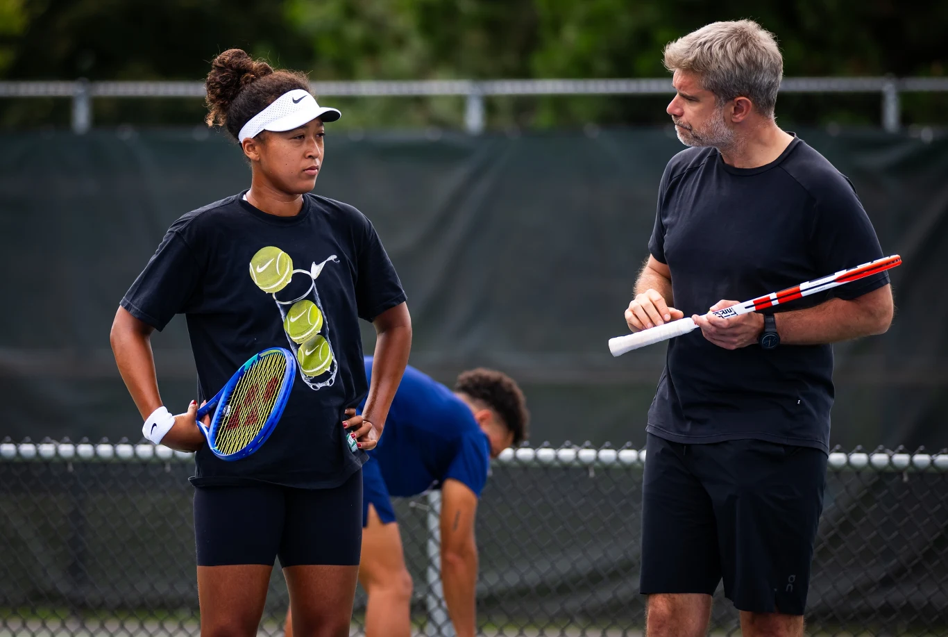 Naomi Osaka i Tomasz Wiktorowski Młoda kobieta w sportowej odzieży i daszku trzymająca rakietę tenisową rozmawia z mężczyzną, który również trzyma rakietę. Obaj stoją na korcie tenisowym, w tle siatka i inna ćwicząca osoba.
