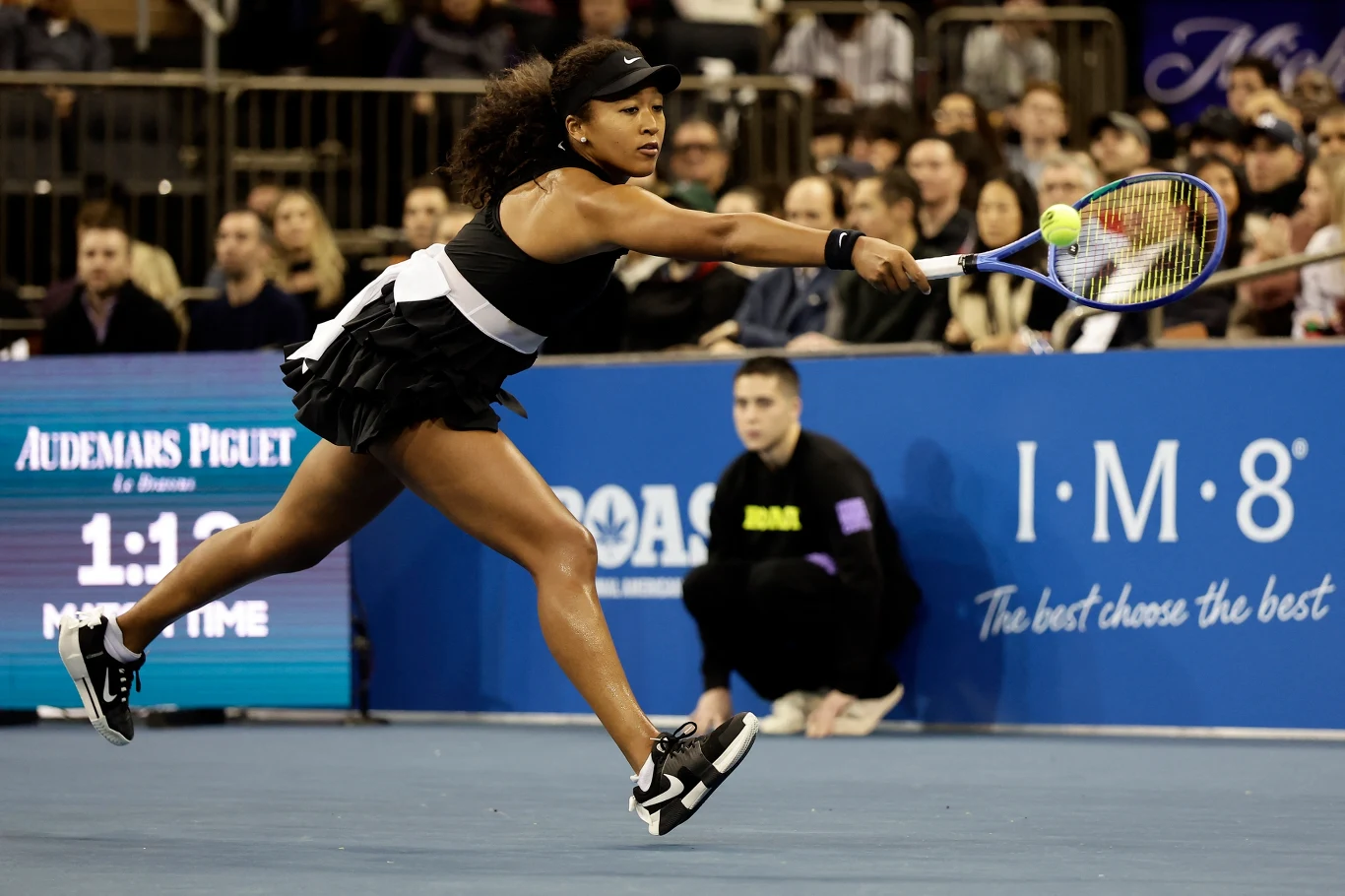 Naomi Osaka Zawodniczka tenisa w czarnej sukience wykonuje dynamiczny ruch, odbijając piłkę rakietą podczas meczu na korcie. W tle widoczna publiczność oraz sędzia siedzący przy linii bocznej.
