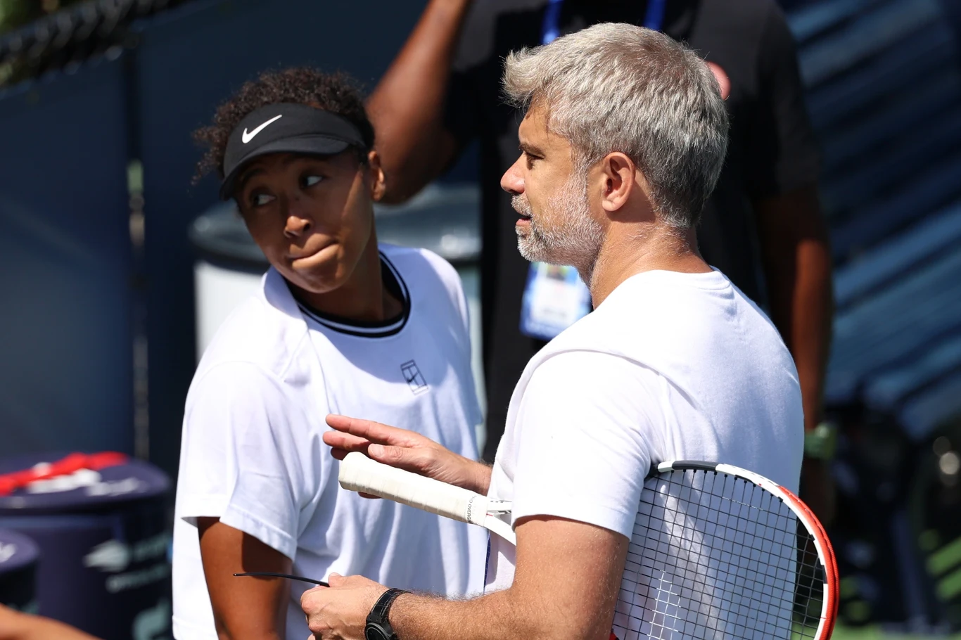 Naomi Osaka i Tomasz Wiktorowski Młoda tenisistka w białej koszulce i czapce z daszkiem rozmawia z trenerem przy korcie tenisowym, trzymającym rakietę; oboje skupieni na rozmowie, otoczeni elementami wyposażenia kortu.