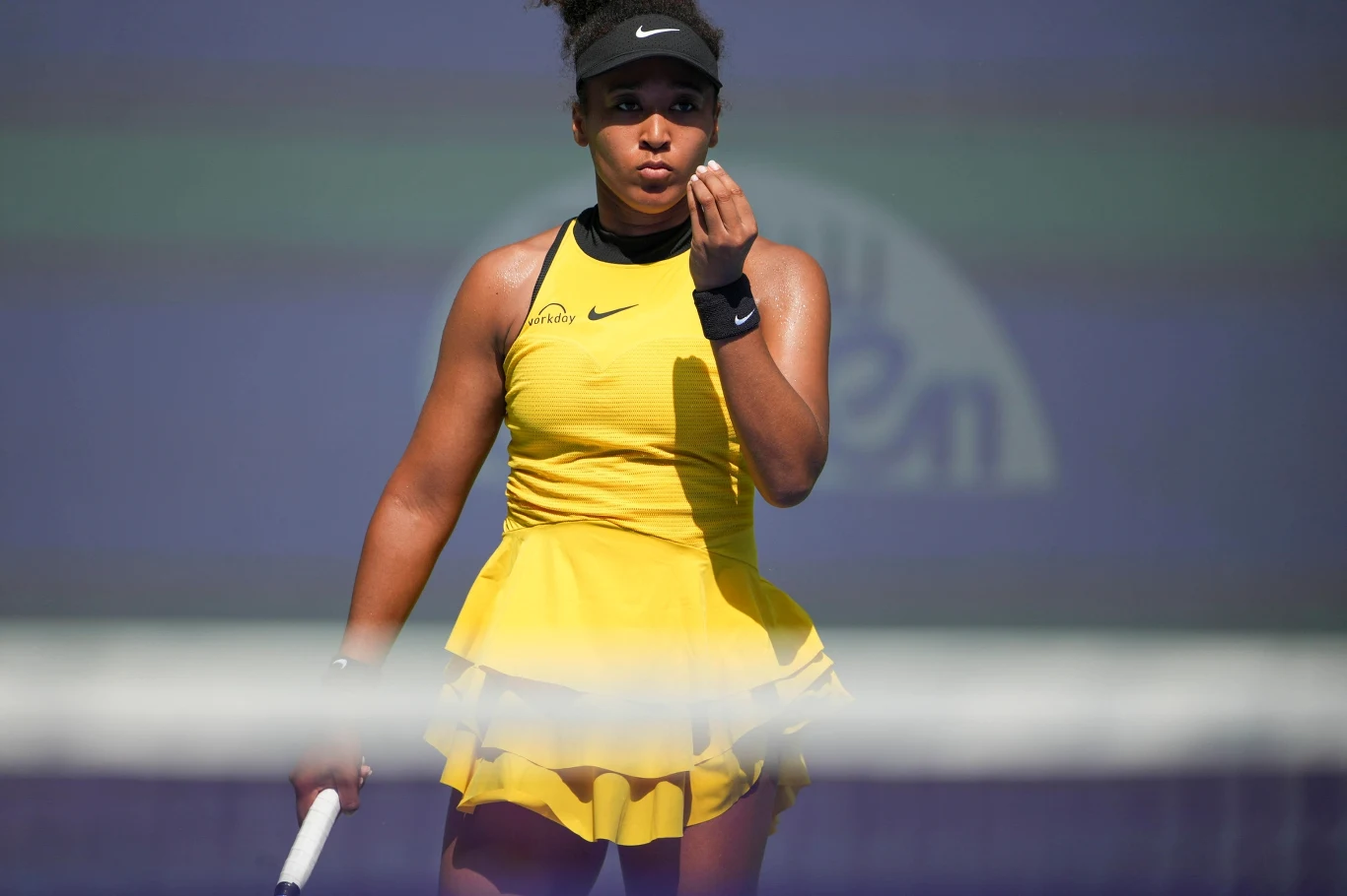 Naomi Osaka Tenisistka ubrana w żółtą sukienkę sportową i czarną czapkę z daszkiem stoi na korcie tenisowym, trzymając rakietę i wykonując gest dłonią w stronę twarzy, co sugeruje skupienie lub analizę sytuacji podczas meczu.