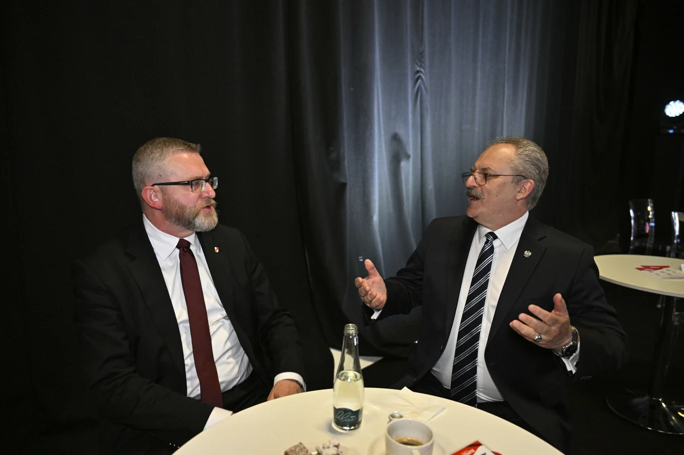 Dwóch mężczyzn w garniturach rozmawiających przy okrągłym stole, na którym znajdują się butelka wody, filiżanka z kawą i talerz. W tle ciemna zasłona, scena oświetlona sztucznym światłem.