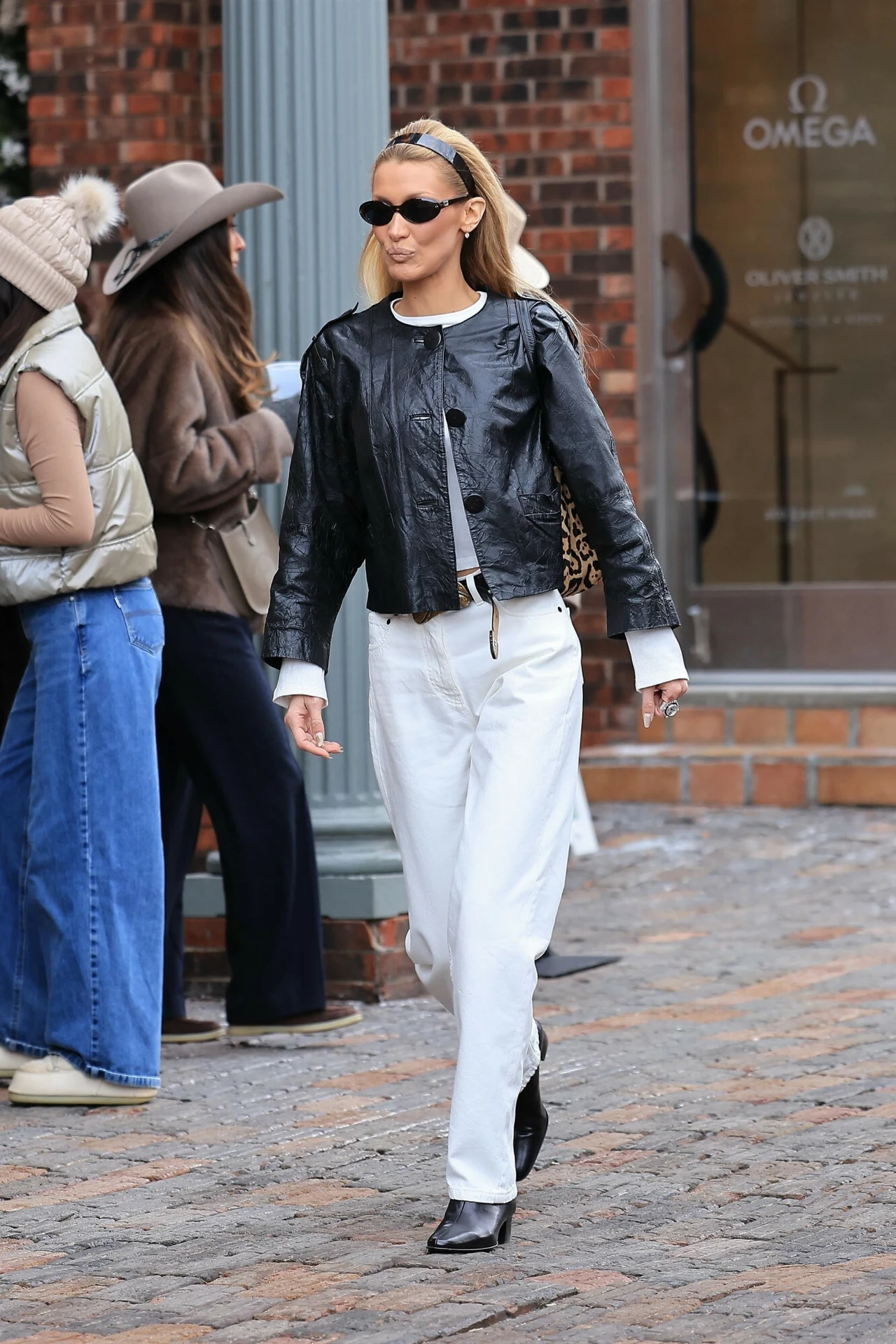 Kobieta ubrana w czarną skórzaną kurtkę, białe spodnie i czarne buty idzie brukowaną ulicą, mając na sobie okulary przeciwsłoneczne oraz opaskę na włosach. W tle grupa osób w luźnych stylizacjach oraz witryna sklepu.