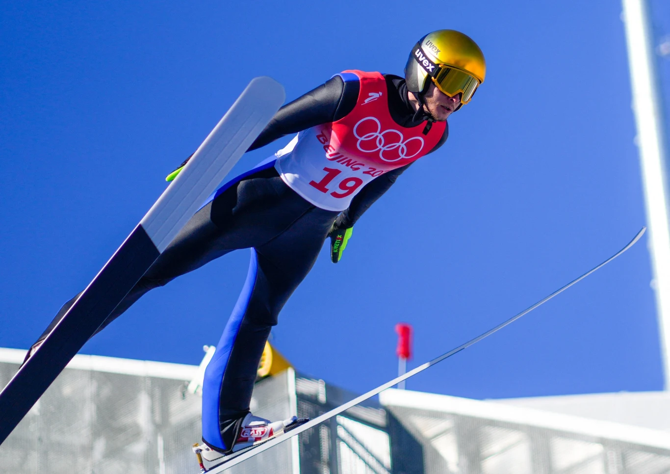 Michaił Nazarow Skoczek narciarski w locie podczas zawodów olimpijskich, ubrany w profesjonalny kombinezon, kask i gogle, z wyraźnym numerem startowym oraz olimpijskim logo na piersi, w tle niebieskie niebo.