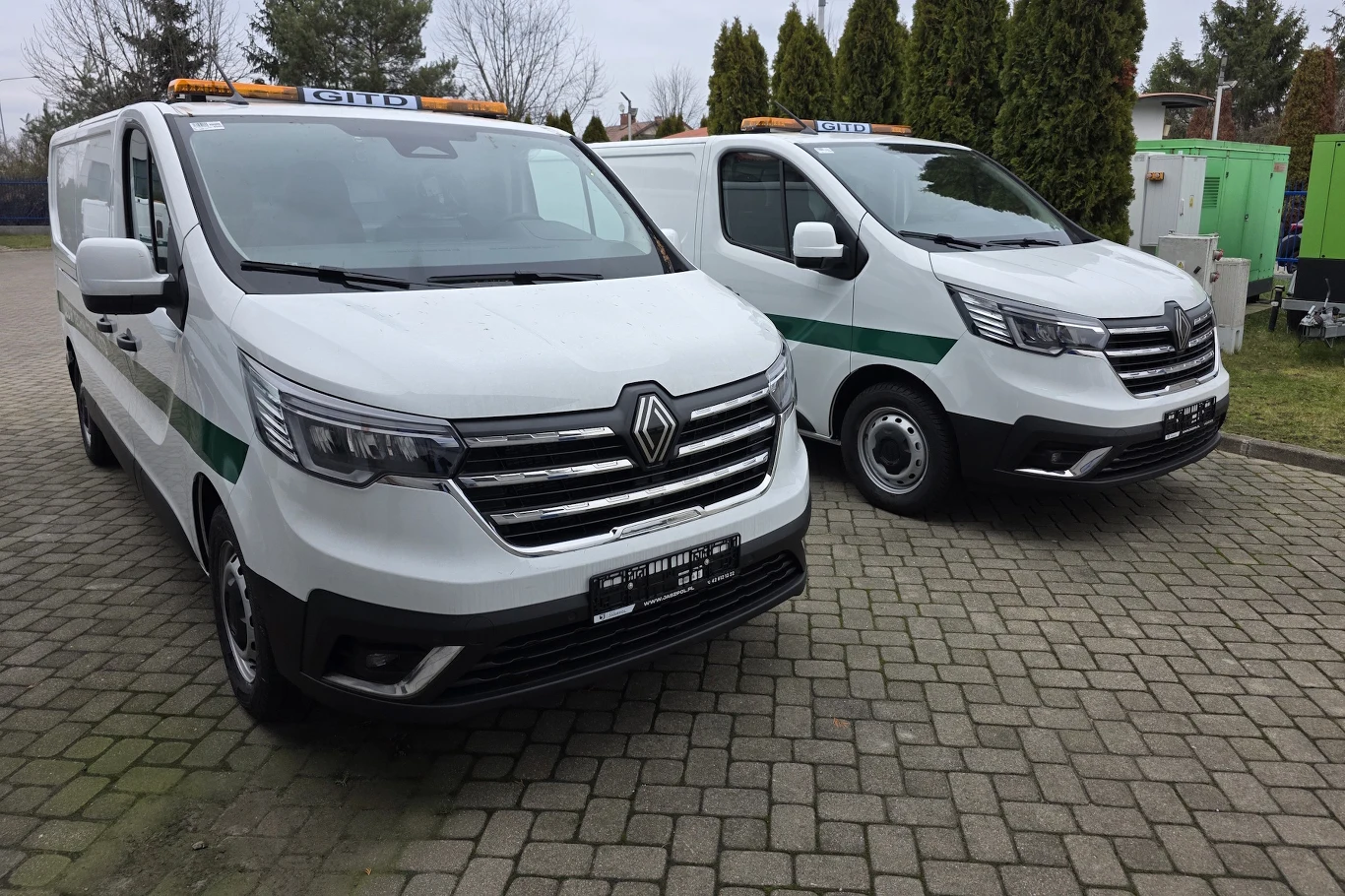 Nowe samochody techniczne GITD Dwa białe samochody Renault z zielonym paskiem oraz oznaczeniem służbowym stoją obok siebie na parkingu z kostki brukowej, w tle widoczne drzewa i elementy infrastruktury.