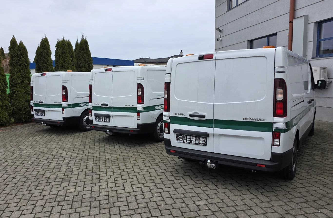 Nowe samochody serwisowe ITD Trzy białe samochody dostawcze marki Renault, należące do służby nadzoru, ustawione równolegle na parkingu przy nowoczesnym budynku, otoczone zielonymi tujami i brukowaną nawierzchnią.