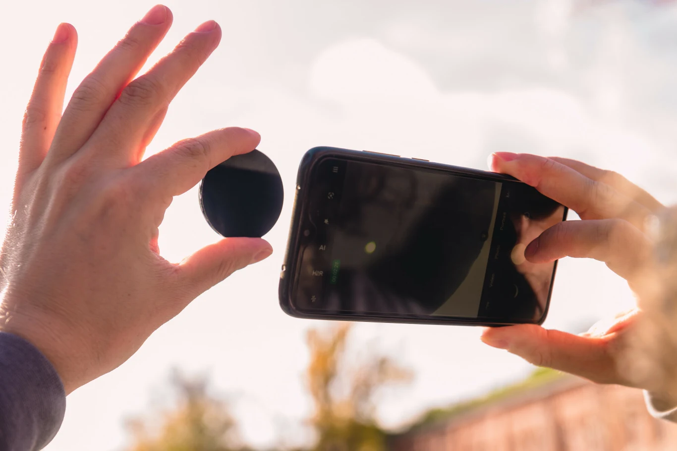 Ręka trzymająca czarny filtr fotograficzny obok smartfona robionego zdjęcia pod światło, z obecnością rozmytego tła i ciepłego światła słonecznego.