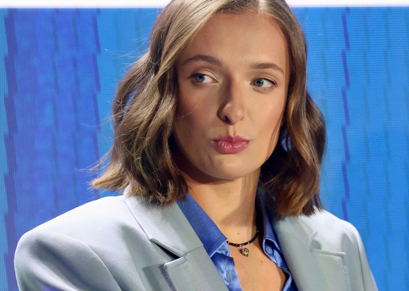 Młoda kobieta z krótkimi, falowanymi włosami, ubrana w jasny garnitur i niebieską koszulę, patrzy w bok z delikatnym uśmiechem. W tle znajduje się niebieski, abstrakcyjny wzór.