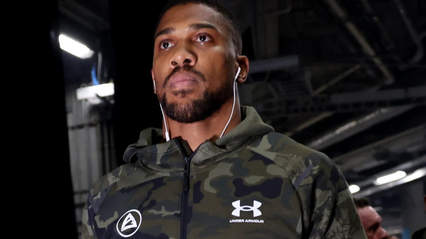 Mężczyzna w sportowej ‌bluzie z zielonym, ⁢militarnym wzorem, słuchawkami ⁢w uszach spaceruje w industrialnym⁣ otoczeniu hali. Twarz ⁤skupiona i poważna. Widoczne logo Under Armour oraz inny znaczek na bluzie.