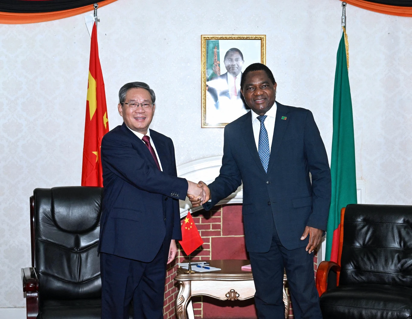 Premier ChRL Li Qiang i prezydent Zambii Hakainde Hichilema podczas spotkania w listopadzie 2025 r. Dwóch mężczyzn w garniturach uśmiecha się i ściska dłonie na tle flag Chin i Zambii, w eleganckim pomieszczeniu z portretem na ścianie.