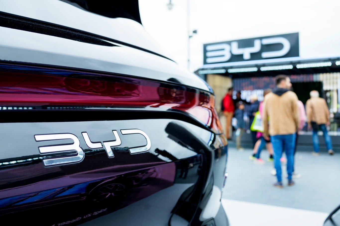 Czarny samochód marki BYD z widocznym logo na tylnej części pojazdu, w tle stoisko wystawowe z logotypem BYD oraz kilka osób przechodzących obok.