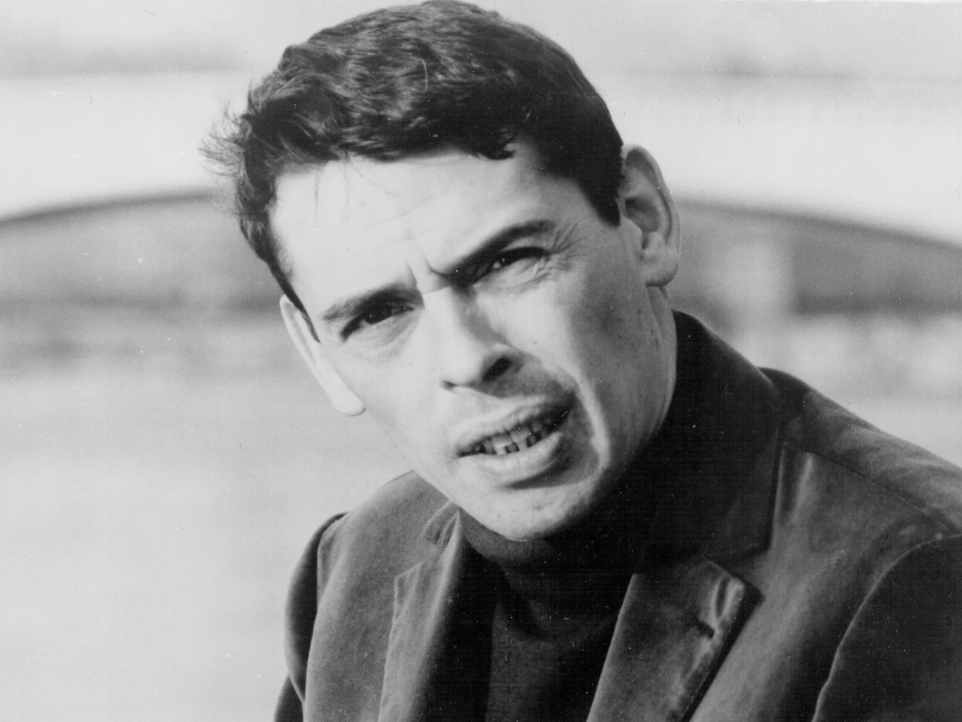 Jacques Brel stworzył utwór "Ne me quitte pas" Mężczyzna o ciemnych włosach i wyrazistych rysach twarzy, ubrany w ciemny golf oraz marynarkę, spogląda w stronę obiektywu na tle rozmytej scenerii z elementem mostu.