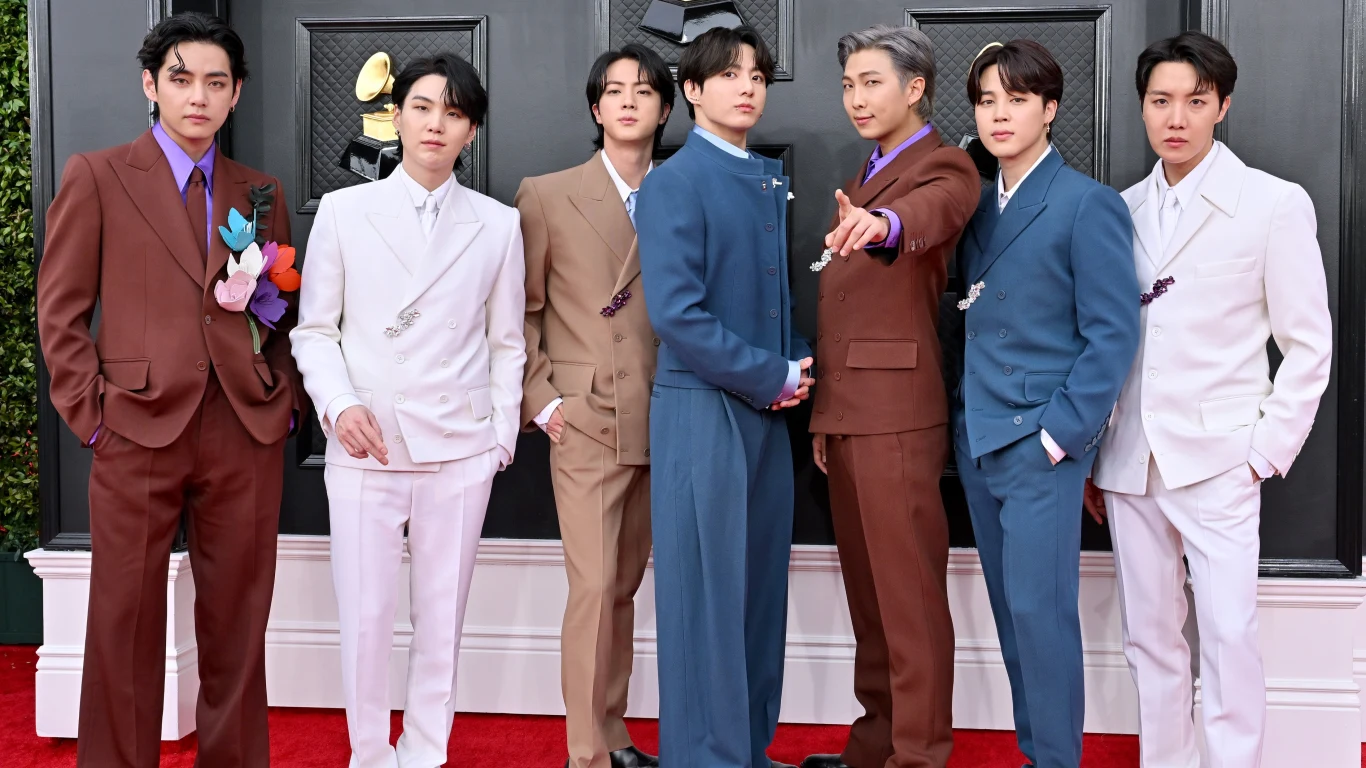BTS, od lewej: V, Suga, Jin, Jungkook, RM, Jimin i J-Hope Siedmiu młodych mężczyzn w eleganckich garniturach pozuje wspólnie na czerwonym dywanie, tworząc zwartą grupę przed oficjalnym tłem eventowym. Każdy ma inny garnitur w różnych odcieniach brązu, niebieskiego, bieli i beżu, wyróżniając się indywidualnym ...