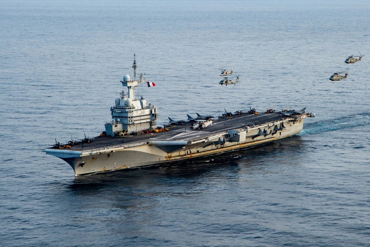 Lotniskowiec wojenny płynący po otwartym morzu z widocznymi na pokładzie samolotami bojowymi. Nad wodą leci kilka wojskowych helikopterów. Na szczycie masztu powiewa flaga Francji.