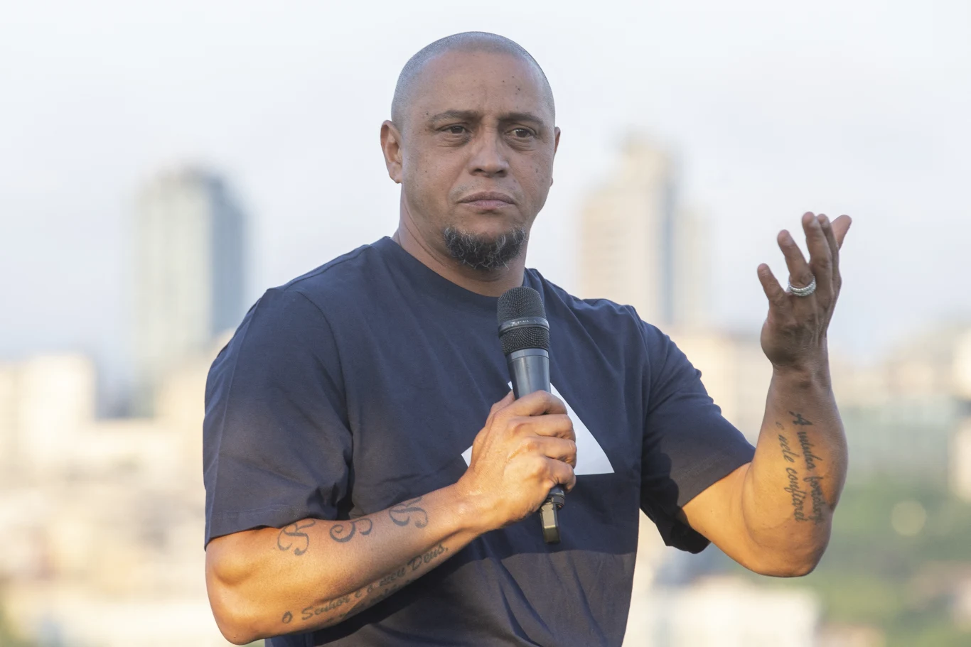 Mężczyzna z krótkimi włosami i tatuażami na obu przedramionach trzyma mikrofon, gestykulując jedną dłonią, stoi na tle rozmytych budynków w oddali.