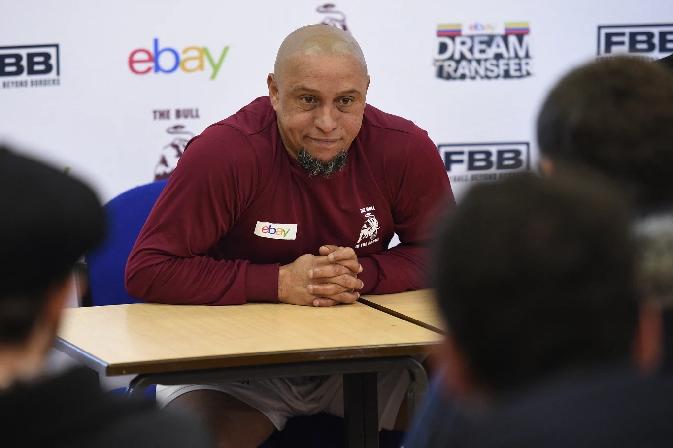 Łysy mężczyzna z brodą siedzi przy stole na konferencji prasowej, ubrany w bordową bluzę sportową, za nim ścianka z logotypami marek oraz kilku obserwatorów zwróconych w jego stronę.