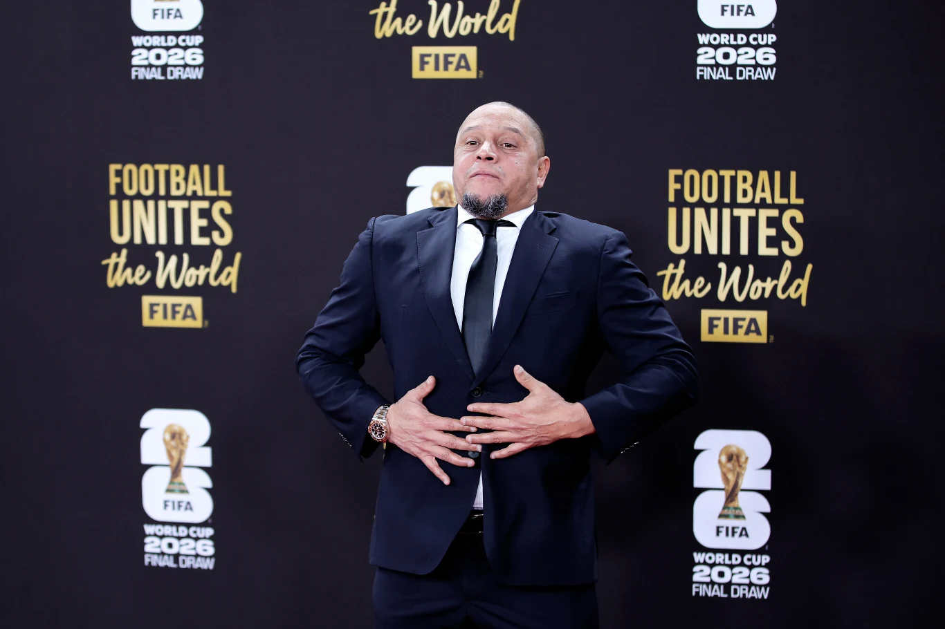 Mężczyzna ubrany w czarny garnitur i białą koszulę pozuje na tle ścianki z logotypami FIFA oraz hasłem Football Unites the World.