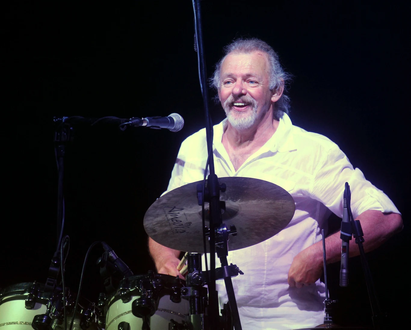 Jerzy Skrzypczyk - najstarszy, wciąż działający na scenie, perkusista w Polsce Starszy mężczyzna z siwą brodą i włosami grający na perkusji podczas koncertu, uśmiechnięty, ubrany w białą koszulę, w tle ciemne tło podkreślające sylwetkę artysty.