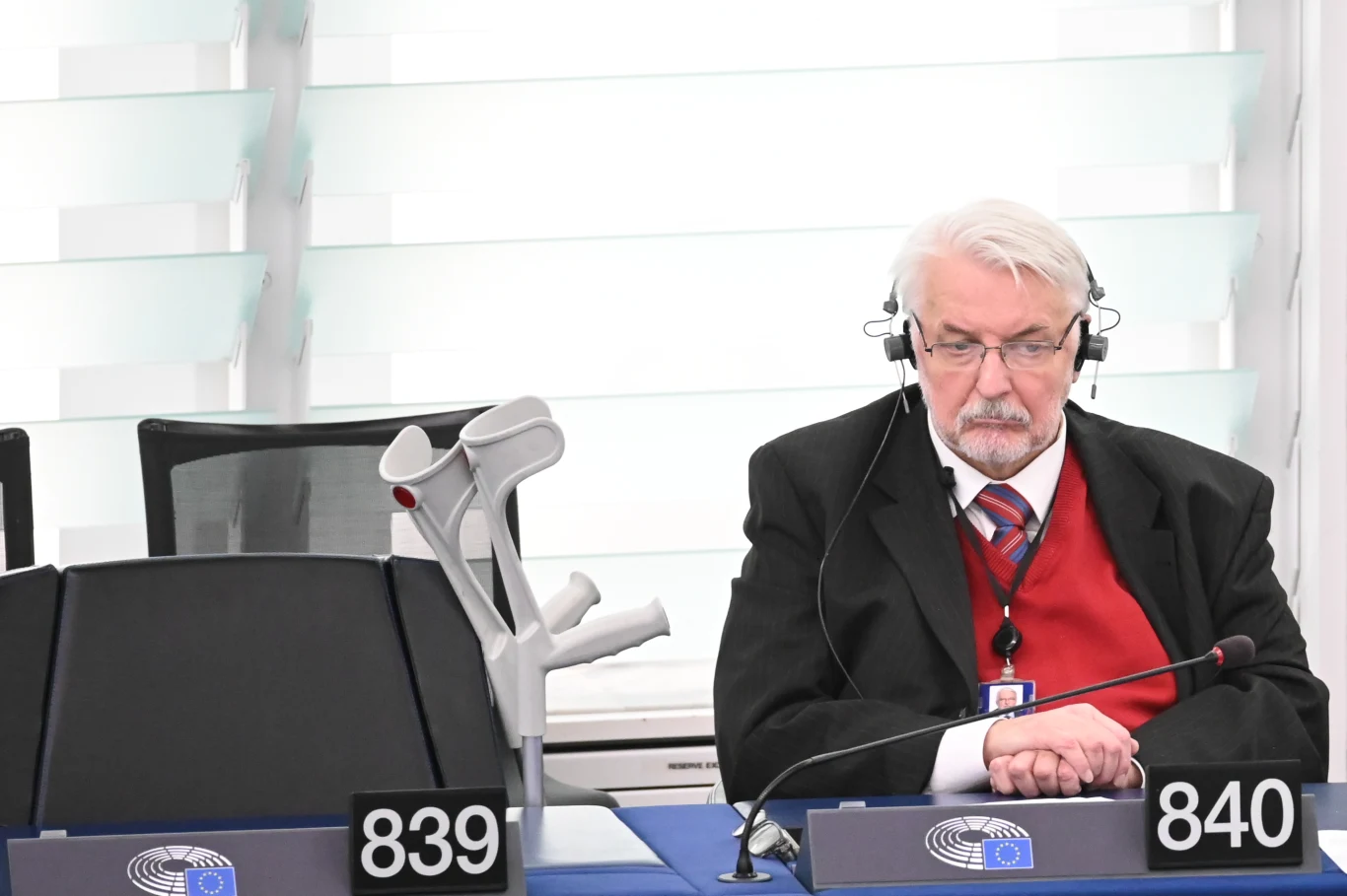 Witold Waszczykowski w europarlamencie 15 lutego 2023 roku Starszy mężczyzna z białymi włosami i brodą siedzi przy stanowisku oznaczonym numerami 839 i 840 w pomieszczeniu urzędowym, nosi garnitur z czerwonym swetrem i słuchawkami na uszach, obok oparte kule ortopedyczne.
