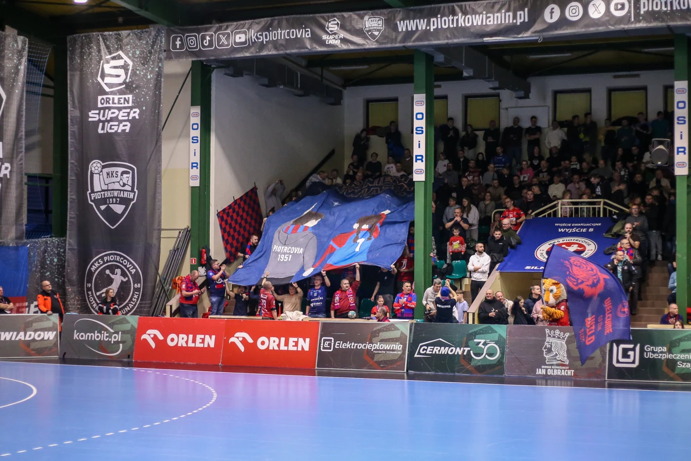 Tłum kibiców na trybunach hali sportowej skandujący i prezentujący duże transparenty, otoczony banerami z logo sponsorów oraz lig sportowych, na pierwszym planie fragment niebieskiego boiska.