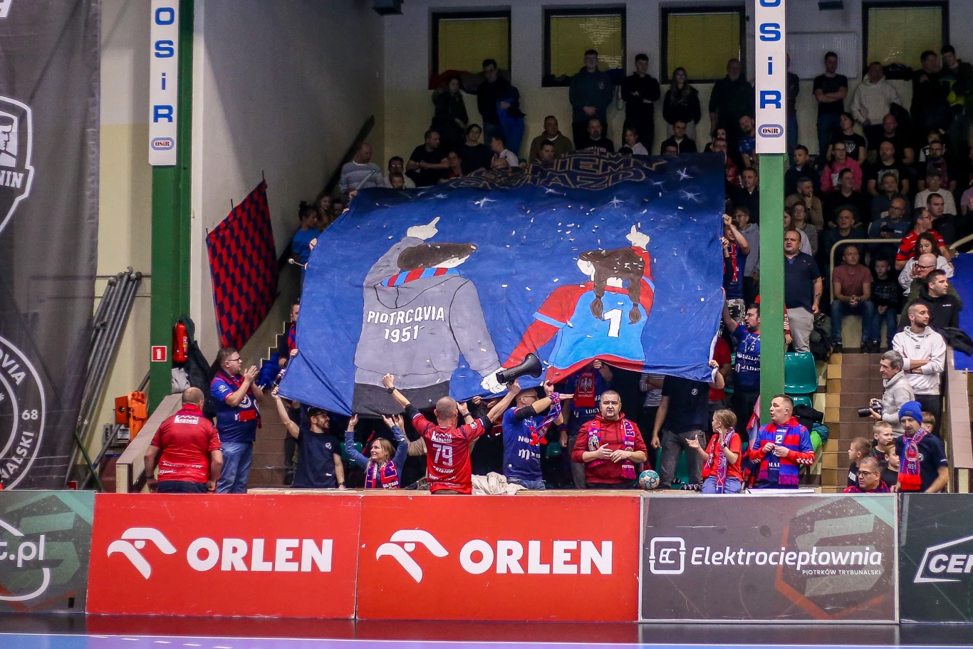 Grupa kibiców ubranych w klubowe barwy na trybunach podnosi duży transparent z grafiką symbolizującą drużynę Piotrcovia 1951. W tle widzowie obserwują wydarzenia sportowe. Na barierkach widoczne są reklamy sponsorów, głównie firmy Orlen.