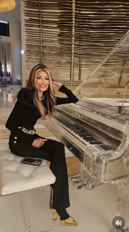 Kobieta z długimi blond włosami w eleganckim, czarnym stroju z ozdobnym paskiem siedzi na białej pufie przy przezroczystym fortepianie, trzymając rękę na instrumencie. Przed nią leży telefon, a w tle widoczne są unikalne dekoracje w złotych barwach.