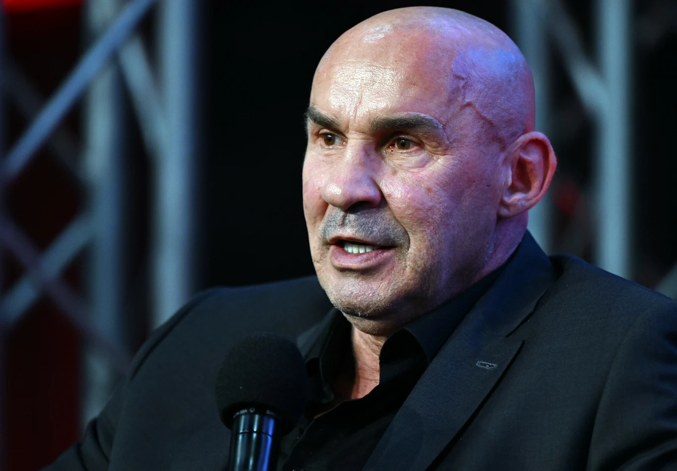 Łysy mężczyzna w garniturze trzyma mikrofon i mówi, tło stanowią metalowe elementy konstrukcyjne oświetlone światłem studyjnym.